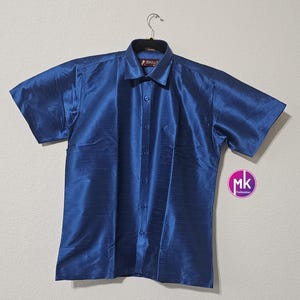 May include: A blue short-sleeved button-down shirt with a subtle sheen. The shirt has a collar and a button closure. The brand name "Angelic" is visible on the collar.