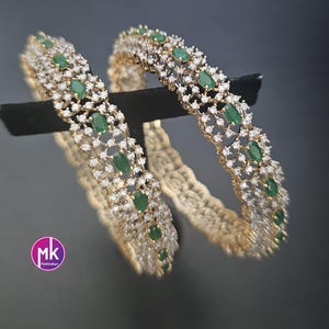 May include: Two gold-tone bangles featuring a lattice design encrusted with small clear stones and oval-shaped green stones. The bangles have a metallic finish and a circular shape. A small circular logo with the letters MK and the text FashionKart is visible.