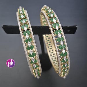 May include: Two gold-tone bangles featuring a central row of alternating oval green stones and marquise-cut clear stones, bordered by thin lines of small clear pavé stones. The jewelry displays a circular design with a metallic finish.