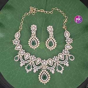 May include: A gold-toned necklace and matching earrings set. The jewelry features clear and emerald-green gemstones in a floral and teardrop design. The set is displayed on a green leaf.