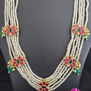 May include: A multi-strand pearl necklace with five strands of white pearls. Each strand has a gold pendant with pink and green gemstones. The necklace is on a black background.