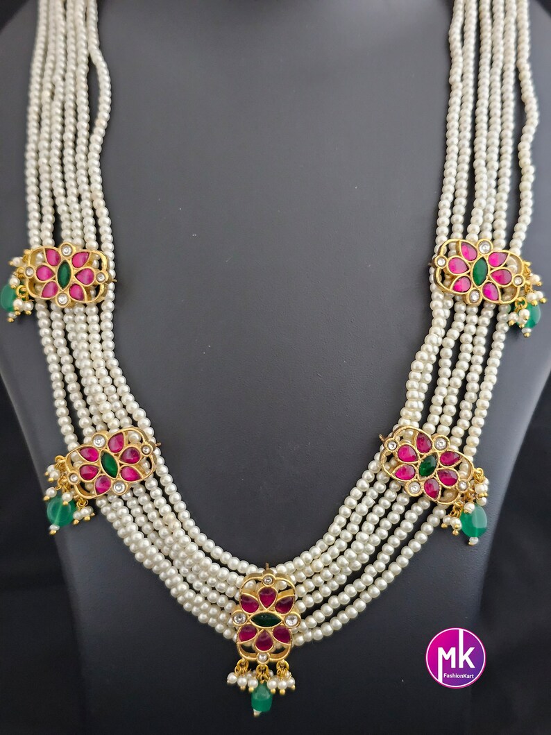 May include: A multi-strand pearl necklace with five strands of white pearls. Each strand has a gold pendant with pink and green gemstones. The necklace is on a black background.