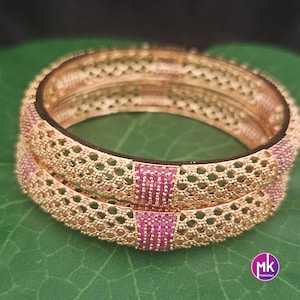 May include: Two gold-colored bangles with intricate openwork and pink accents. The bangles are stacked, showcasing a detailed design with small, evenly spaced holes and rectangular pink sections. The background is a green surface.