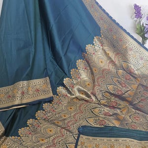 Puede incluir: Un sari de seda azul turquesa con un borde dorado y negro. El borde presenta intrincados dise&ntilde;os florales y paisley. El sari tiene un patr&oacute;n tejido de peque&ntilde;os puntos en el pallu.