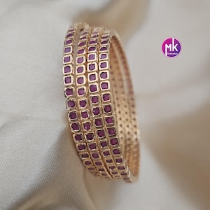 May include: A set of three gold-toned bangles featuring a continuous row of square-cut, deep magenta-colored stones set within each band. The metal surface displays a textured, perforated pattern along the sides, and a circular logo with the text mk.