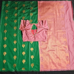 May include: A green and pink silk saree with a gold tree pattern on the green side and a gold geometric pattern on the pink side. The saree is draped over a table and a pink blouse is laid on top of the saree.