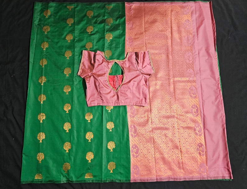 May include: A green and pink silk saree with a gold tree pattern on the green side and a gold geometric pattern on the pink side. The saree is draped over a table and a pink blouse is laid on top of the saree.