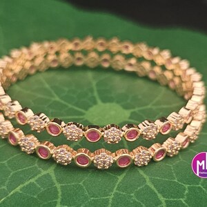 May include: Two gold-colored bangles with alternating red and clear gemstones. The bangles are set on a green leaf. The gemstones are round and set in a floral pattern. The bangles are likely made of metal.