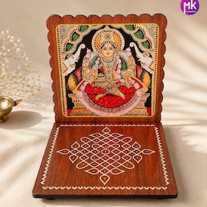 May include: A wooden folding stand featuring a painted illustration of a deity seated on a lotus, flanked by two elephants. The base displays a white geometric kolam pattern. The top edge has a scalloped design, and a logo reads mk FashionKart.