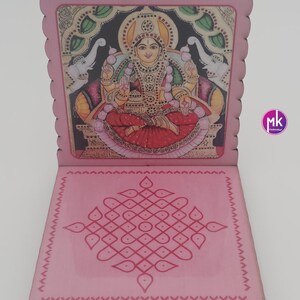 May include: A pink wooden folding stand featuring a scalloped edge. The upright panel displays a colorful illustration of a deity seated on a throne with two elephants. The base panel features a symmetrical pink geometric kolam pattern design.