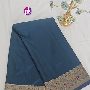 Puede incluir: Un sari azul turquesa con un borde dorado y marr&oacute;n con un patr&oacute;n floral. El sari est&aacute; doblado y colocado plano sobre una superficie blanca.