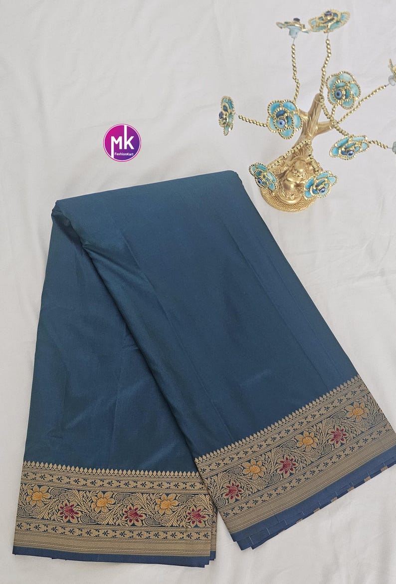 Puede incluir: Un sari azul turquesa con un borde dorado y marr&oacute;n con un patr&oacute;n floral. El sari est&aacute; doblado y colocado plano sobre una superficie blanca.