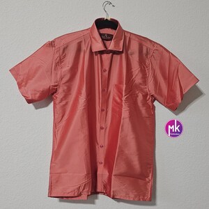 May include: A coral colored short-sleeved button-down shirt with a pocket on the left side. The shirt is made of a silky material and has a collar.