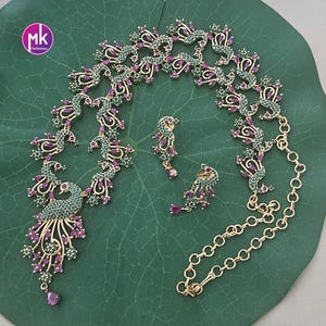 May include: A gold-coloured necklace and earring set featuring a peacock design. The necklace and earrings are adorned with green and purple gemstones. The jewellery is displayed on a green leaf-shaped surface.
