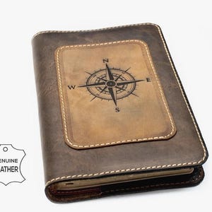 Könnte beinhalten: Ein dunkelbraunes Lederjournal mit einem rechteckigen, hellbraunen Ledereinsatz auf dem Vorderdeckel. Der Einsatz ist mit einer schwarzen Kompassrose und den Buchstaben N, E, S und W geprägt. Ein weißes, lederförmiges Etikett zeigt GENUINE LEATHER.