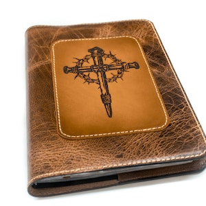 Puede incluir: Un diario de cuero marrón con un diseño de cruz en relieve. La cruz, hecha de clavos y una corona de espinas, está sobre un parche de cuero marrón claro. El diario tiene un borde cosido y una apariencia texturizada.