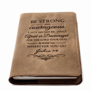 Puede incluir: Un diario de cuero color canela con el texto "BE STRONG and courageous" y un versículo bíblico de Josué 1:9. La cubierta tiene un borde cosido y un lomo marrón oscuro. El diario está cerrado y parece nuevo.