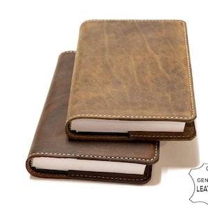 May include: Two brown leather journals with white stitching and blank pages. The top journal is slightly larger than the bottom journal. The journals are stacked on top of each other.