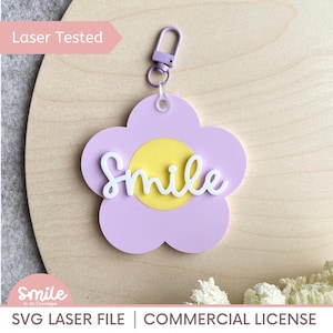Może przedstawiać: Brelok w kształcie kwiatu w kolorze liliowym z żółtym środkiem i napisem "Smile" w białym skrypcie. Posiada srebrny zatrzask. Tekst "Laser Tested" znajduje się w lewym górnym rogu. Radosny dodatek.
