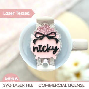 Pode incluir: Um topper de copo personalizado rosa e brilhante com o nome "nicky" em escrita preta. O topper tem um laço preto e é preso a um copo branco. As palavras "Laser Tested" estão no canto superior esquerdo.