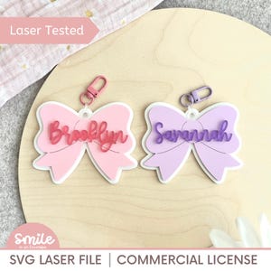 Può includere: Due portachiavi a forma di fiocco, uno rosa e uno lavanda, ciascuno con un nome in corsivo. Il portachiavi rosa dice "Brooklyn" e il portachiavi lavanda dice "Savannah". Entrambi hanno un contorno bianco e un fermaglio in metallo.