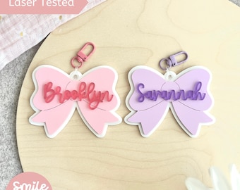 May include: Two bow-shaped keychains, one pink and one lavender, each with a name in a cursive font. The pink keychain says "Brooklyn" and the lavender keychain says "Savannah". Both have a white outline and a metal clasp.