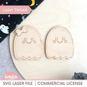 Op de afbeelding: Twee houten spookuitsparingen met lachende gezichten en geschulpte bodems. Eén spook heeft een strik. De tekst "Laser Tested" staat in een roze doos. De tekst "SVG LASER FILE | COMMERCIAL LICENSE" staat onderaan. De achtergrond is licht hout.