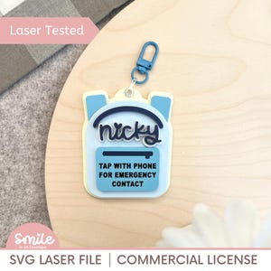 May include: A light blue backpack-shaped keychain with the name "nicky" in white script. The keychain has a blue carabiner clip and includes the text "TAP WITH PHONE FOR EMERGENCY CONTACT". The words "Laser Tested" are in the upper left corner.