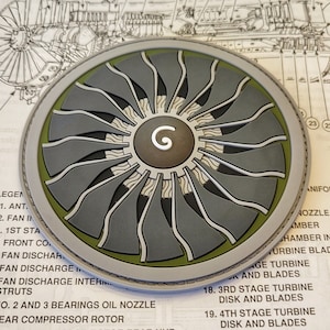 May include: A circular metal emblem featuring a detailed illustration of a jet engine fan. The fan blades are gray and white, set against a dark gray background. The center has a circular design with the letter "G". The emblem is on a technical diagram.