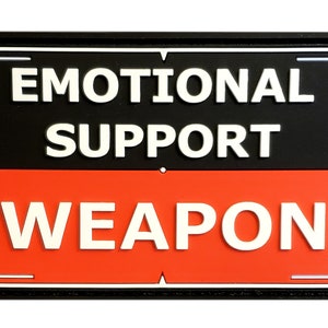 May include: A rectangular patch with the words "EMOTIONAL SUPPORT WEAPON" in white, bold letters. The top half is black, and the bottom half is red. The patch has a black border.