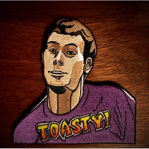 Peut inclure: Écusson brodé représentant un homme de dessin animé aux cheveux bruns et une chemise violette. La chemise porte le mot "TOASTY!" en lettres orange et jaune. L'écusson est posé sur un fond en bois.