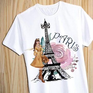 Puede incluir: Camiseta blanca con un estampado gr&aacute;fico de la Torre Eiffel, una mujer con un vestido marr&oacute;n y la palabra "PAR&Iacute;S" en letras negras. El gr&aacute;fico est&aacute; rodeado de flores de acuarela rosa y verde.