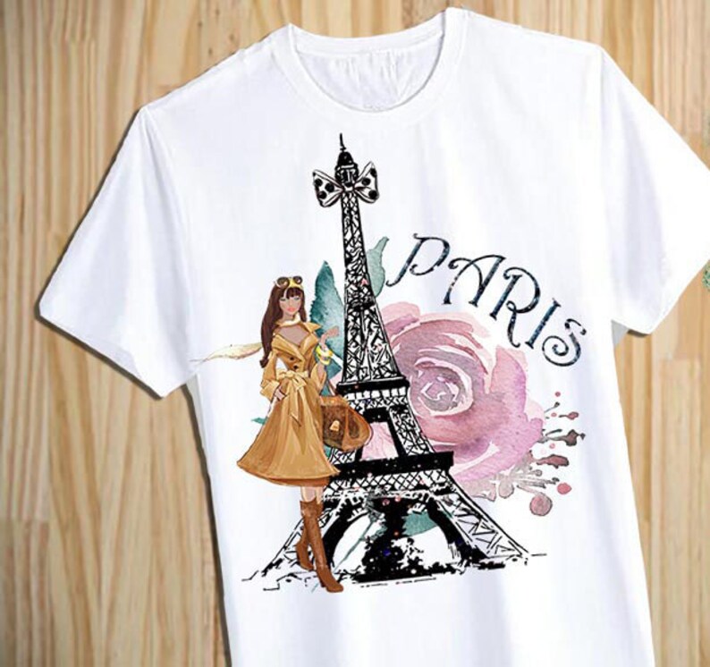 Puede incluir: Camiseta blanca con un estampado gr&aacute;fico de la Torre Eiffel, una mujer con un vestido marr&oacute;n y la palabra "PAR&Iacute;S" en letras negras. El gr&aacute;fico est&aacute; rodeado de flores de acuarela rosa y verde.