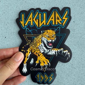 May include: A die-cut sticker featuring a snarling yellow and black spotted jaguar leaping forward. The design includes the text JAGUARS in yellow, Cosmic Disco in white, and 1995 in yellow, set against a black background with blue lightning.