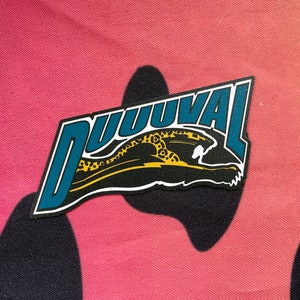 May include: A patch featuring the word "DUUUVAL" in teal letters with a white outline. The patch includes a black and gold jaguar graphic. The background is a pink and black patterned fabric.