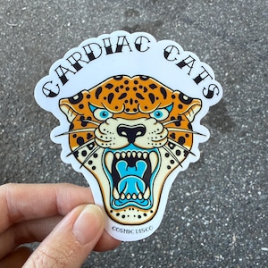 May include: A sticker featuring a roaring jaguar with orange spots, blue eyes, and a turquoise mouth. The sticker has the text "CARDIAC CATS" in a curved font above the jaguar and "COSMIC DISCO" below.