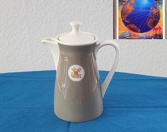 Könnte beinhalten: Eine grau-weiße Keramik-Kaffeekanne mit Deckel und Griff. Der Topf hat ein goldenes Emblem auf der Vorderseite. Im Hintergrund befindet sich ein Vintage-Reiseposter mit einem Globus und den Worten "Norddeutscher Lloyd Bremen."