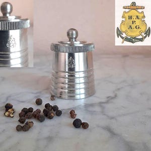 May include: A silver-toned metal pepper shaker with a ribbed cylindrical body and a matching lid featuring a small spherical knob. The side of the shaker is embossed with a crest displaying the letters H.A.P.A.G. above an anchor symbol.