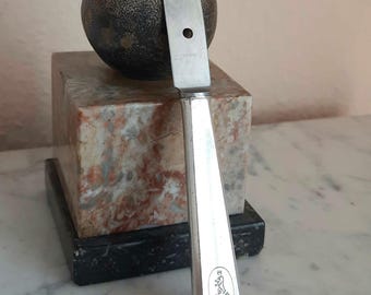Könnte beinhalten: Ein silberfarbenes Metallmesser mit flachem, spitz zulaufendem Griff, das ein ovales Bärenemblem und den Schriftzug INOX auf der Klinge aufweist. Das Messer steht vertikal vor einer dunklen, texturierten Kugel auf einem quadratischen Marmorsockel.