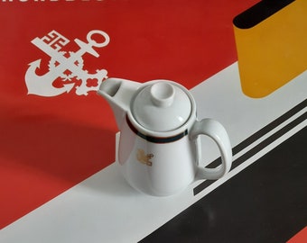 Könnte beinhalten: Kleine, weiße Keramik-Teekanne mit Deckel, Ausguss und Henkel. Die Teekanne hat ein goldenes Emblem und ein grün-schwarzes Band am oberen Rand. Der Hintergrund zeigt den Text "NORDDEUTSCHER LLOYD" und ein rot-weiß-schwarz-gelbes Design.