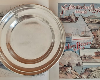 Könnte beinhalten: Ein silberner Teller mit einem erhöhten Rand und einem kleinen Logo in der Mitte. Der Teller liegt auf einem Buch mit einem Vintage-Reiseposter für Norddeutscher Lloyd Bremen, das Bilder von Italien und den Text "Italien Reisen" zeigt.