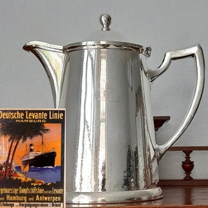 Peut inclure: Une cafetière argentée brillante avec un couvercle et une poignée. La cafetière a un design classique et un petit emblème. Une affiche de voyage vintage pour Deutsche Levante Linie, Hambourg, est visible.