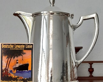 Könnte beinhalten: Eine glänzende silberne Kaffeekanne mit Deckel und Griff. Die Kanne hat ein klassisches Design und ein kleines Emblem. Ein Vintage-Reiseposter für Deutsche Levante Linie, Hamburg, ist sichtbar.