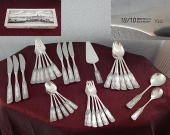 Könnte beinhalten: Eine Sammlung silberfarbener Besteckteile, darunter Messer, Gabeln, Löffel und ein Tortenheber, angeordnet auf einer weinroten Oberfläche. Das Besteck hat dekorative Griffe. Eine Schachtel mit einem Schwarz-Weiß-Landschaftsbild befindet sich in der oberen linken Ecke. Das Besteck ist mit "18/10 Solingen Germany 100" gekennzeichnet.