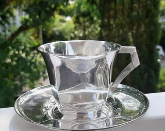 Könnte beinhalten: Ein glänzendes silbernes Tee-Tassen- und Untertassen-Set. Die Tasse hat einen geometrischen Griff und einen ausgestellten Rand, der auf einer passenden Untertasse steht. Das Set ist hochreflektierend, mit einem Logo auf der Tasse. Der Hintergrund ist verschwommenes grünes Laub.