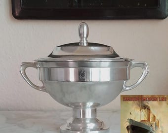 Könnte beinhalten: Eine silberfarbene Metallsuppenterrine mit Deckel, zwei seitlichen Griffen und einem Standfuß. Der Körper zeigt ein kleines eingraviertes Emblem. Daneben befindet sich ein rechteckiges Bild eines großen Schiffes mit dem Text HAMBURG-AMERICAN LINE.