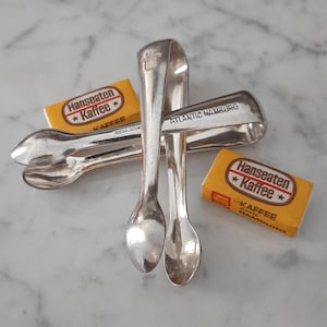 May include: A pair of silver-toned metal sugar tongs with the text ATLANTIC HAMBURG engraved on one side, positioned alongside two small rectangular yellow boxes labeled Hanseaten Kaffee with a star graphic and additional text.