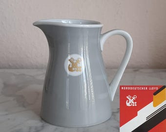 Könnte beinhalten: Graue Keramikkrug mit weißem Innenraum und Griff. Ein goldenes Emblem befindet sich auf der Vorderseite. Ein Vintage-Reiseposter für Bremen New York befindet sich im Hintergrund.