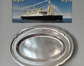 Könnte beinhalten: Eine ovale Servierplatte aus silberfarbenem Metall mit gewelltem Rand und einer glatten, reflektierenden Oberfläche. Über der Platte befindet sich ein rechteckiges Poster mit einem großen Ozeandampfer auf See und dem Text United American Lines.
