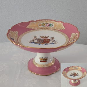 Op de afbeelding: Een porseleinen schaal op voet met een roze rand voorzien van gouden filigraanaccenten en een witte kom in het midden versierd met een kleurrijk heraldisch wapen. De voet is wit met een roze rand en een kroonmotief, afgewerkt met goud.